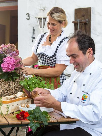Ein Koch bereitet frische Zutaten vor, während eine Frau im traditionellen Trachtenkleid Blumen arrangiert. Im Hintergrund sind Kräuter und Gemüse auf einem Tisch verteilt.