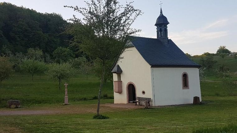 Eine kleine Kapelle in einer grünen Landschaft, umgeben von Bäumen. Der Himmel ist klar und die Atmosphäre ist ruhig.