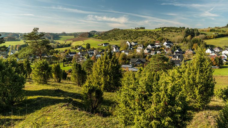 Eine malerische Landschaft mit einem kleinen Dorf, umgeben von Hügeln und Wäldern. Der Himmel ist klar und die Sonne scheint.