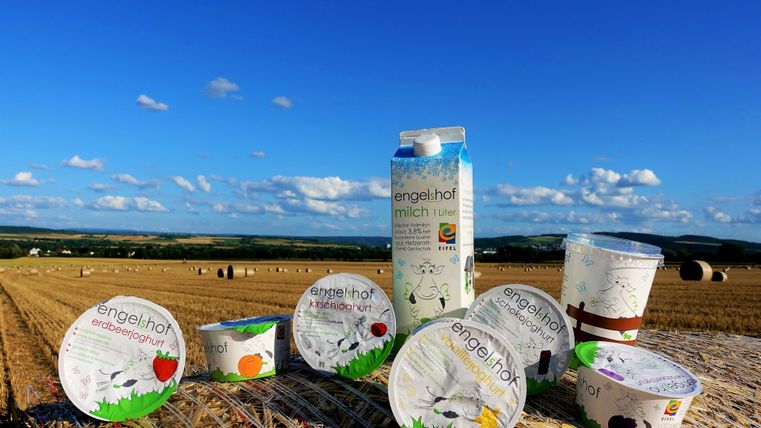 Eine Auswahl an Molkereiprodukten von Engelhof steht auf einem Feld bei blauem Himmel. Im Hintergrund sind strohballen und eine weite Landschaft zu sehen.