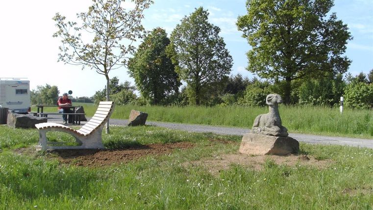 Eine grüne Wiese mit Bäumen und einer Sitzbank. Im Hintergrund steht eine Statue und eine Person sitzt an einem Tisch.