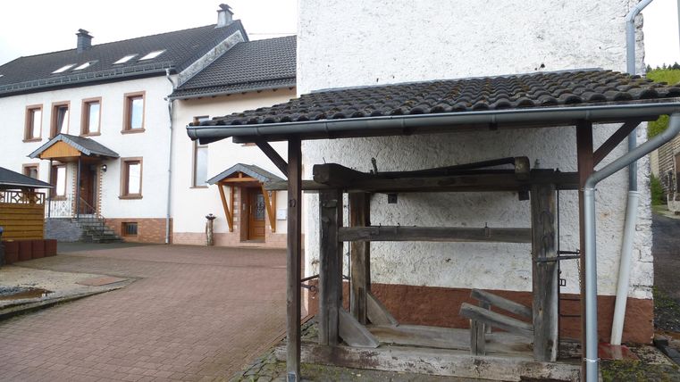 Ein einfaches Gebäude mit weißen Wänden und Holzverkleidungen. Im Vordergrund steht eine alte Holzstruktur.