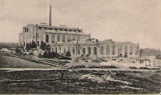 Een historisch fabrieksgebouw met grote ramen en een schoorsteen. Het bevindt zich in een landelijke omgeving met enkele bouwplaatsen en onverharde oppervlakken.