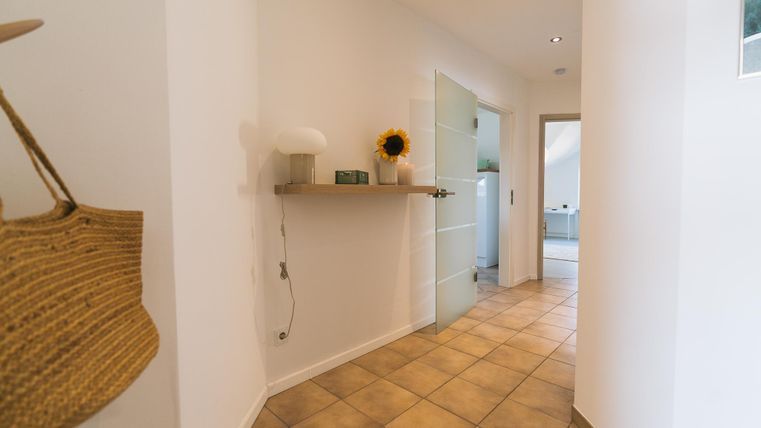 Een lichte gang met lichte wanden en een betegelde vloer. Op een plank staat een lamp en een zonnebloem.