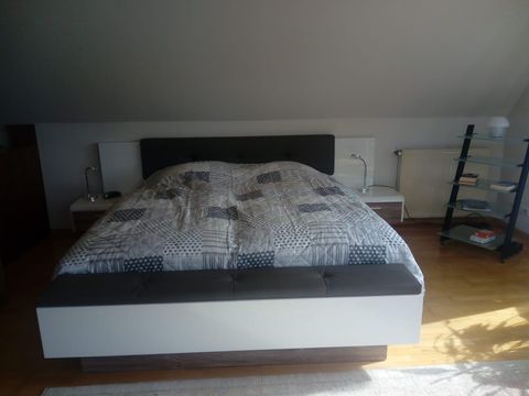 Een moderne slaapkamer met een groot bed en een gezellige deken. Daarnaast staat een glazen nachtkastje en een subtiel verlichte lamp.