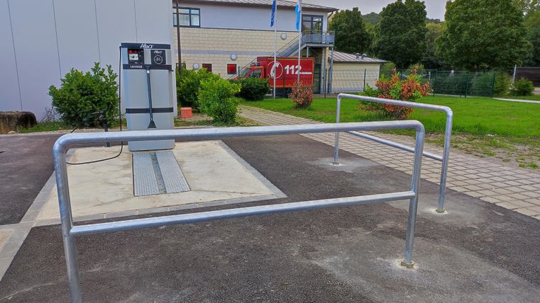 A modern gas station with a pumping station and a fenced-off area. In the background, buildings and trees can be seen.