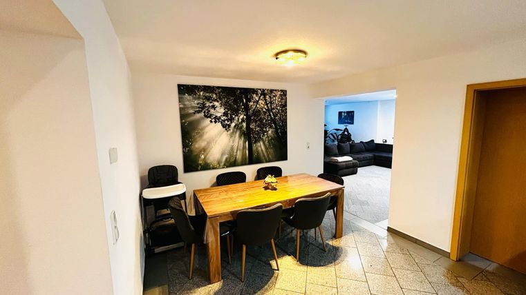 A modern dining area with a wooden table and black chairs. A picture of sun rays through trees hangs on the wall.