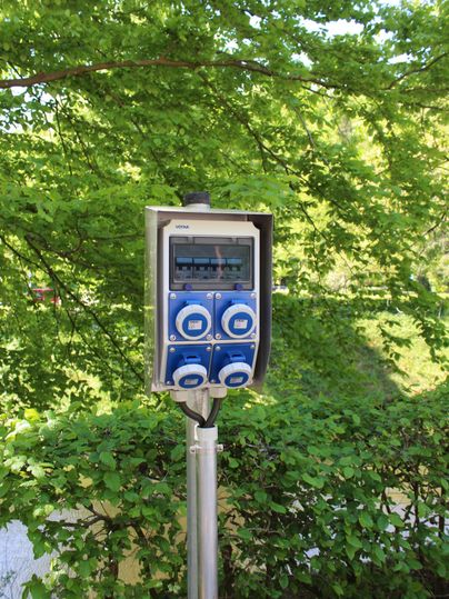 Ein elektrischer Zählerkasten steht auf einem Stahlpfosten, umgeben von grünem Laub. Die Schaltbox hat blaue Bedienelemente und ist im Freien installiert.