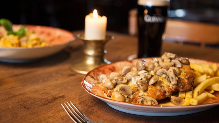 Ein leckerer Teller mit gebratenem Fleisch, überzogen mit Pilzen und serviert mit Pommes. Im Hintergrund gibt es eine Kerze und ein Pint Bier.
