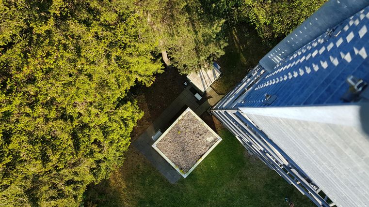 A bird's-eye view of a garden with a small pavilion and surrounding trees. In the background, a high-rise building can be seen.