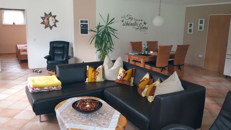 A cozy living room with a large sofa corner and decorative cushions. In the background, a dining table and a plant are visible.