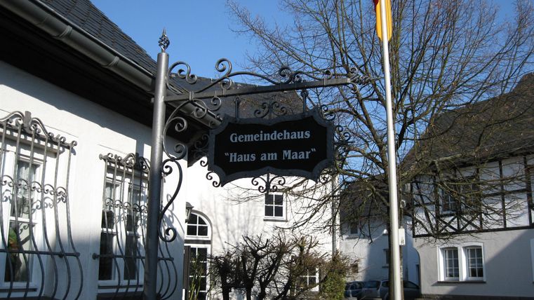Ein hübsches Gebäude mit einem historischen Schild. Es zeigt das Gemeindehaus "Haus am Maar" und ist von einem klaren blauen Himmel umgeben.