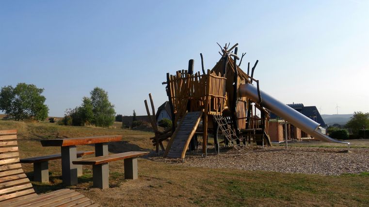Ein Spielplatz aus Holz mit Rutschen und Kletterstrukturen. Im Vordergrund sind eine Bank und ein Tisch zu sehen.