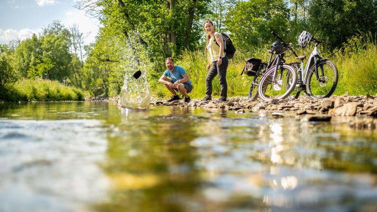 Zwei Personen stehen am Ufer eines Flusses und werfen Steine ins Wasser. Neben ihnen stehen zwei Fahrräder. Die Umgebung ist grün und sonnig.