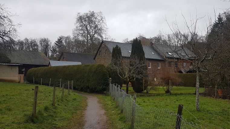 Ein malerischer Weg führt durch eine grüne Wiese zu einem historischen Bauernhaus. Im Hintergrund sind Bäume und weitere Gebäude sichtbar.