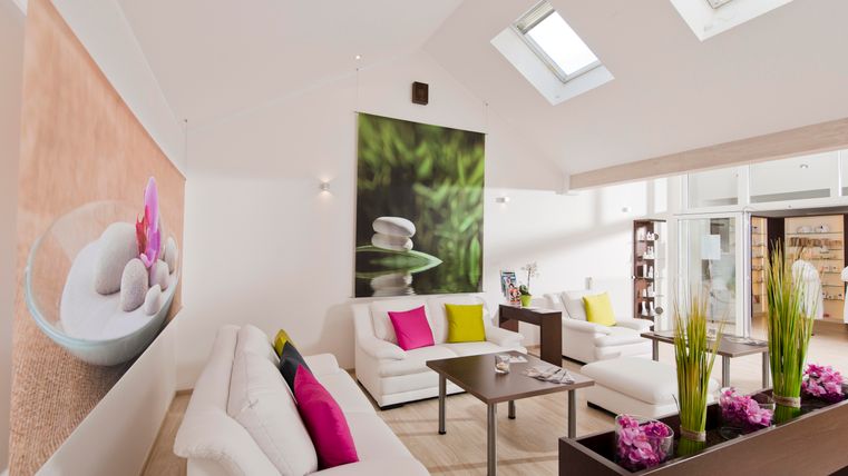 Ein modernes Wohnzimmer mit weißen Sofas und bunten Kissen. Große Fenster sorgen für viel Tageslicht und eine entspannte Atmosphäre.