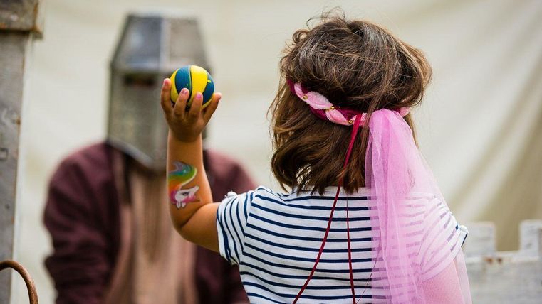 Een kind draagt een kleurrijke doek en houdt een bal in de hand. Op de achtergrond is een persoon in een harnas te zien.