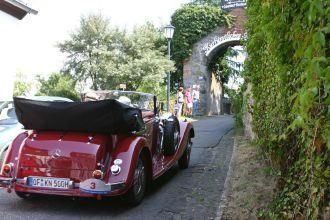 A red vintage convertible is parked on a cobblestone road. In the background, a gate with green vegetation is visible.