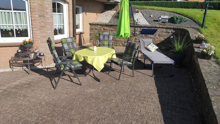 Eine gemütliche Terrasse mit einem runden Tisch, Stühlen und einer Liege. Ein grüner Sonnenschirm spendet Schatten und die Umgebung ist gepflegt.