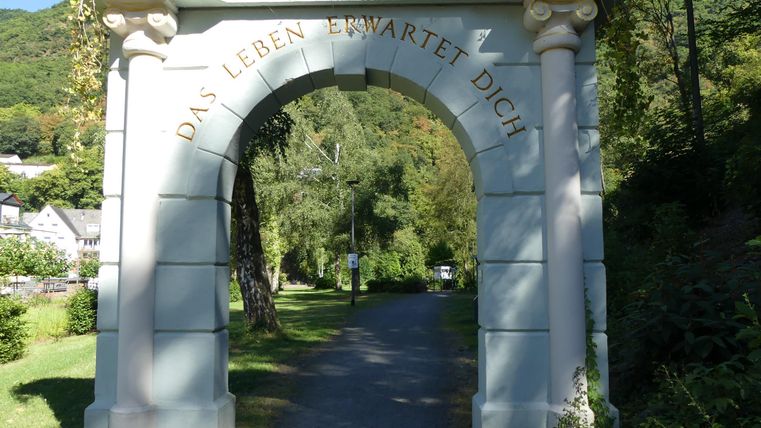 Ein eleganter Torbogen aus Stein mit der Aufschrift "Das Leben wartet auf dich". Der Weg führt durch eine grüne Landschaft mit Bäumen.