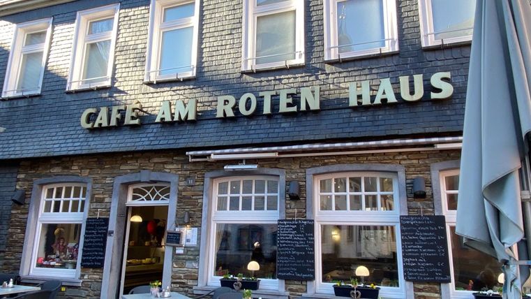 Ein gemütliches Café mit dem Namen „Cafe am Roten Haus“. Es gibt Tische im Freien und eine einladende Atmosphäre.