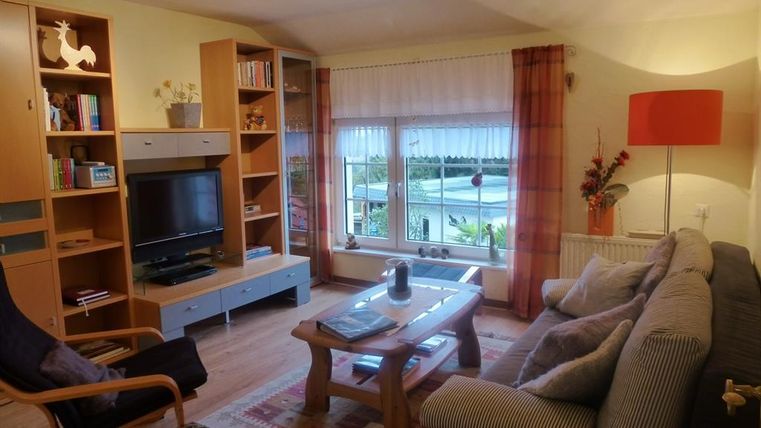 Ein gemütliches Wohnzimmer mit einem großen Sofa und einem Fernseher. Die Fenster bieten einen Blick nach draußen, und es gibt dekorative Elemente wie Pflanzen und eine Lampe.