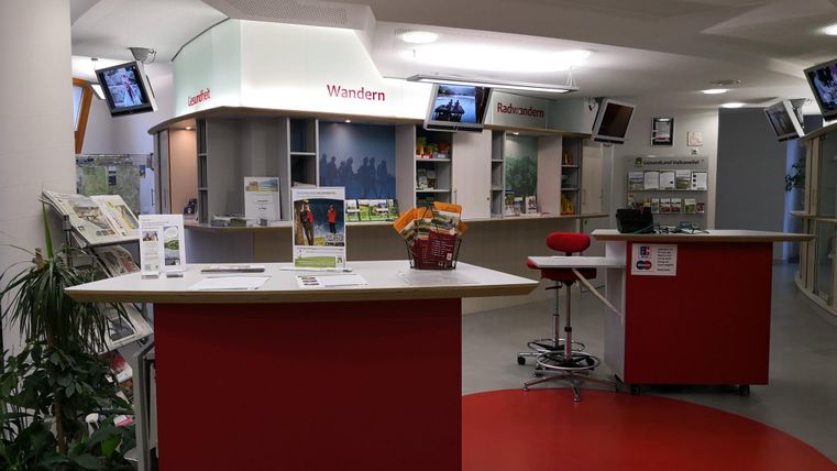 Een modern informatiecentrum met meerdere balies en tv-schermen. De ruimtes zijn helder en vriendelijk ingericht, met planten op de achtergrond.