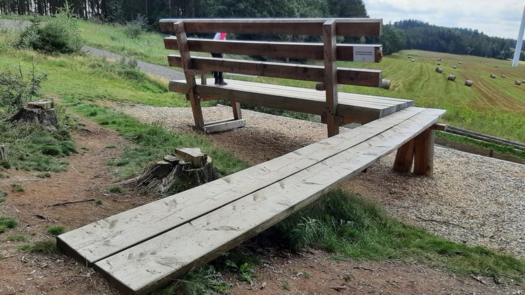 Eine Holzbank mit einer langen Sitzfläche steht in der Natur. Im Hintergrund sind Wiesen und Bäume zu sehen.