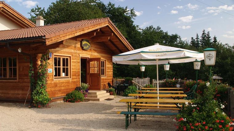 Ein gemütliches Holzhaus mit einer Terrasse und einem großen Sonnenschirm. Vor dem Haus stehen Bänke und Tische unter bunten Blumen.