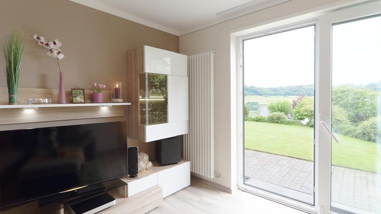 Een moderne woonkamer met een televisie en een grote raampartij. Door het raam is een groene tuin en het landschap zichtbaar.