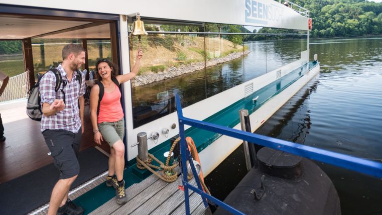 Twee mensen lachen terwijl ze van boord gaan van een modern schip met glazen wanden. Een vrouw luidt een bel. Op de achtergrond is een beboste kust te zien.