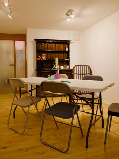 Ein einfacher Raum mit einem Tisch und mehreren Stühlen. Im Hintergrund steht ein antiker Schrank.