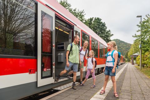A family is getting off a red train at the station. In the background, trees and a green landscape can be seen.