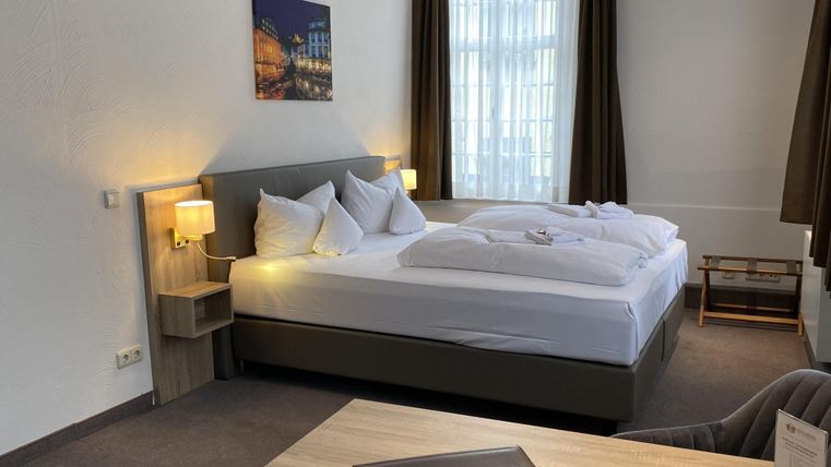 Ein modernes Hotelzimmer mit zwei Betten, Nachttischen und einem Schreibtisch. Helle Vorhänge und ein Bild an der Wand sorgen für eine freundliche Atmosphäre.