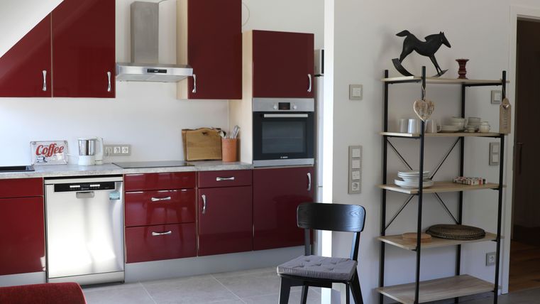 A modern kitchen with red cabinets and white countertops. An open shelf with dishes and decorations enhances the cozy atmosphere.
