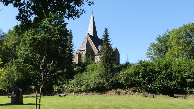 Eine alte Kirche auf einem Hügel, umgeben von Bäumen und grünem Gras. Der Himmel ist klar und blau.
