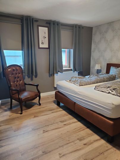 A cozy bedroom with a double bed and an armchair. The walls are brightly decorated, and light streams through the windows.