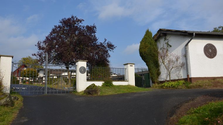 Ein Zugangstor zu einem Grundstück mit einem großen, blühenden Baum. Im Hintergrund sind Gebäude und ein klarer Himmel sichtbar.