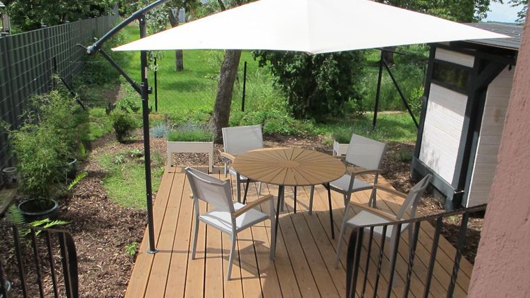 Ein gemütlicher Außenbereich mit einem runden Tisch und vier Stühlen. Ein Sonnenschirm bietet Schatten in einer grünen Umgebung.