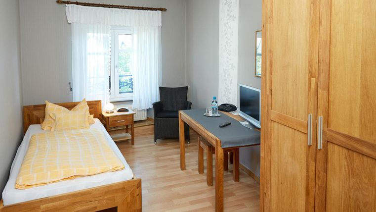 Een eenvoudige kamer met een eenpersoonsbed, een tafel en een stoel. De muren zijn licht en er zijn grote ramen die voor veel licht zorgen.