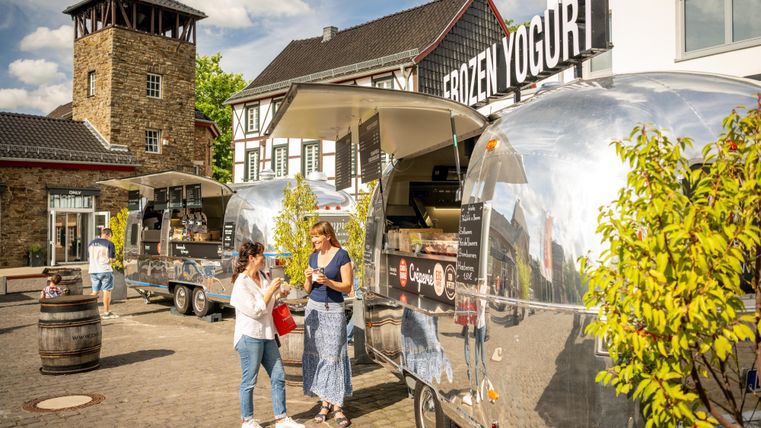 Ein helles und einladendes Szene mit Food Trucks, die gefrorenen Joghurt verkaufen. Zwei Frauen stehen zusammen und genießen die Atmosphäre.