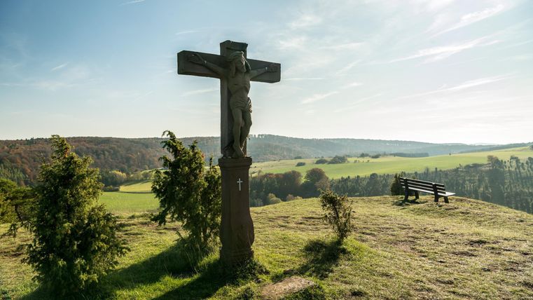 Ein großer Holzkreuz mit einer Statue steht auf einem Hügel. Im Hintergrund sind grüne Wiesen und ein blauer Himmel zu sehen.