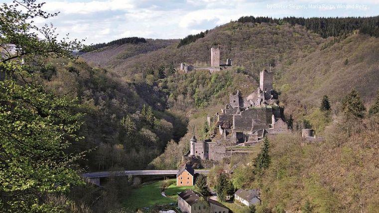 A picturesque landscape with ancient castle ruins on a hill. Green forests and a small village are visible in the foreground.