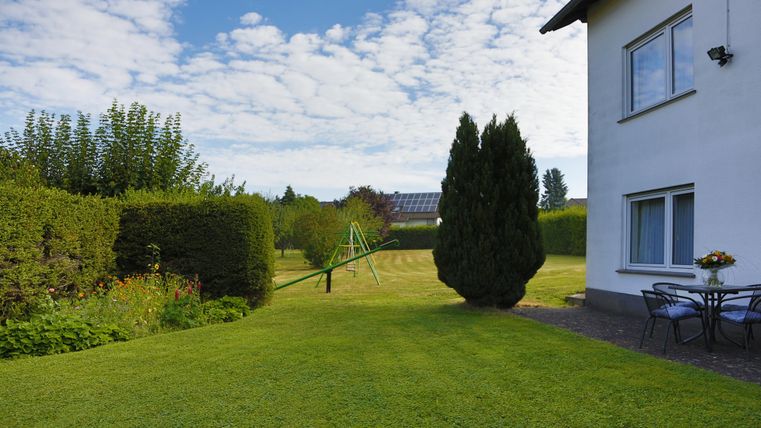 Ein gepflegter Garten mit grünem Rasen und einem Spielplatz im Hintergrund. Am Rand steht ein Haus mit einem schönen, blauen Himmel.