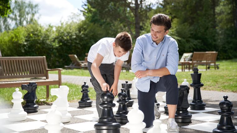 Ein Vater und sein Sohn spielen Schach im Freien auf einem großen Schachbrett. Sie lachen und genießen die Zeit in der Natur.