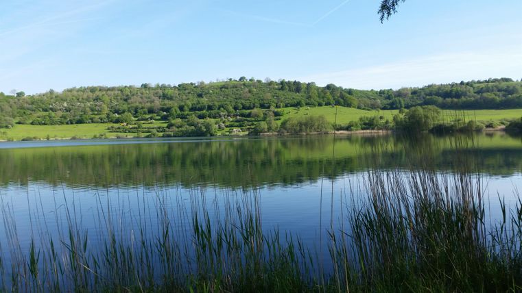 Ein ruhiger See umgeben von üppigem Grün und sanften Hügeln. Die Wasseroberfläche spiegelt die Landschaft wider.