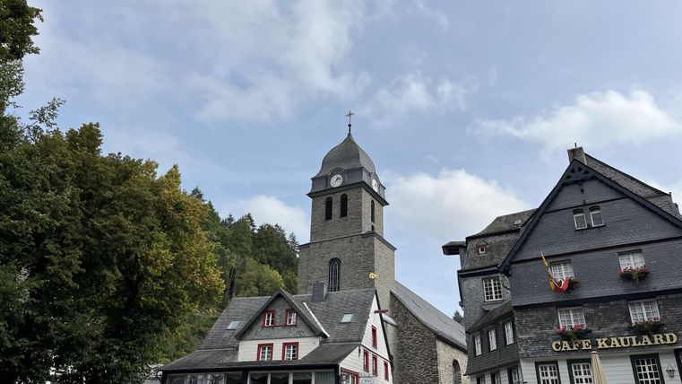 A picturesque square with historic buildings and a striking church tower. The sky is partly cloudy, and there are trees in the background.
