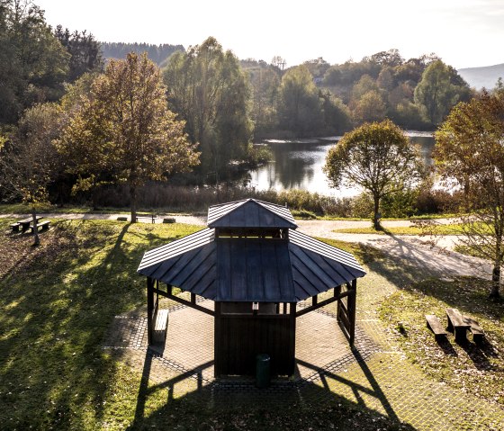Ein Pavillon in einem herbstlichen Park mit B&auml;umen und einem See im Hintergrund. Sonnenlicht wirft lange Schatten auf den Boden., &copy; Eifel Tourismus GmbH, Dominik Ketz