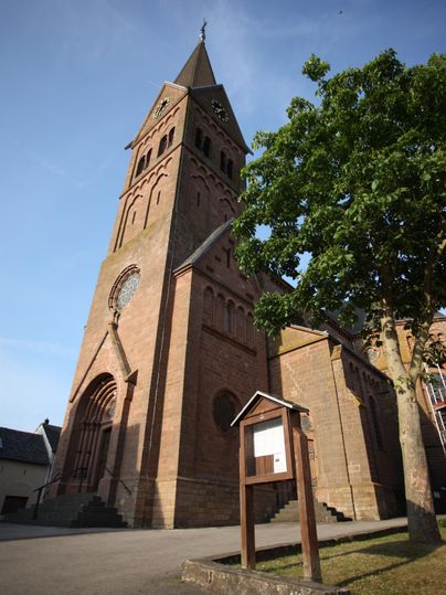 Die Kirche in Niederbettingen mit ihrem hohen Turm aus rotem Stein, daneben ein grüner Baum und ein Schaukasten im Vordergrund.