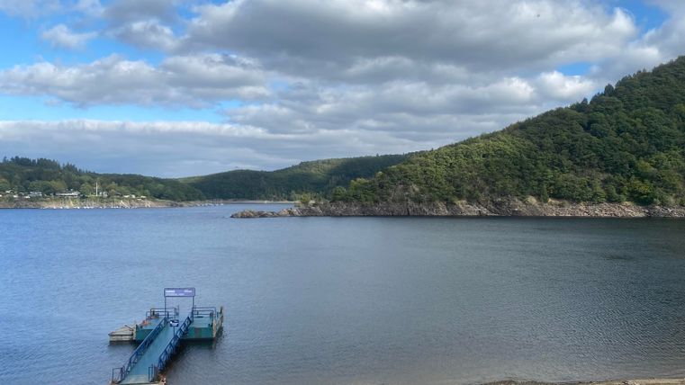 A tranquil lake with a pier and gentle hills in the background. The sky is lightly cloudy and reflects in the water.